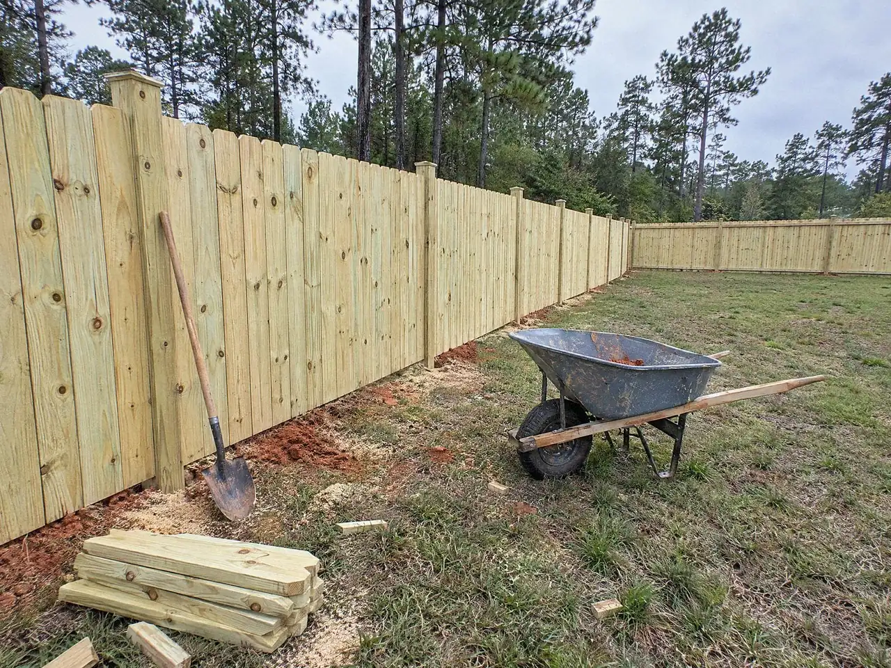 Wood privacy fence line with a shovel leaning on a post and a wheelbarrow on the grass